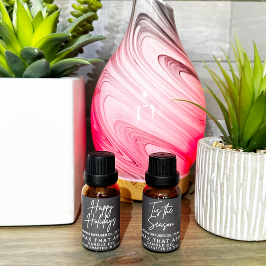 Two essential oil bottles with labels next to a pink and white marbled diffuser with plants in the background.