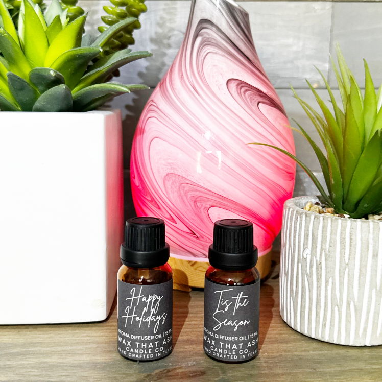 Two essential oil bottles with labels next to a pink and white marbled diffuser with plants in the background.