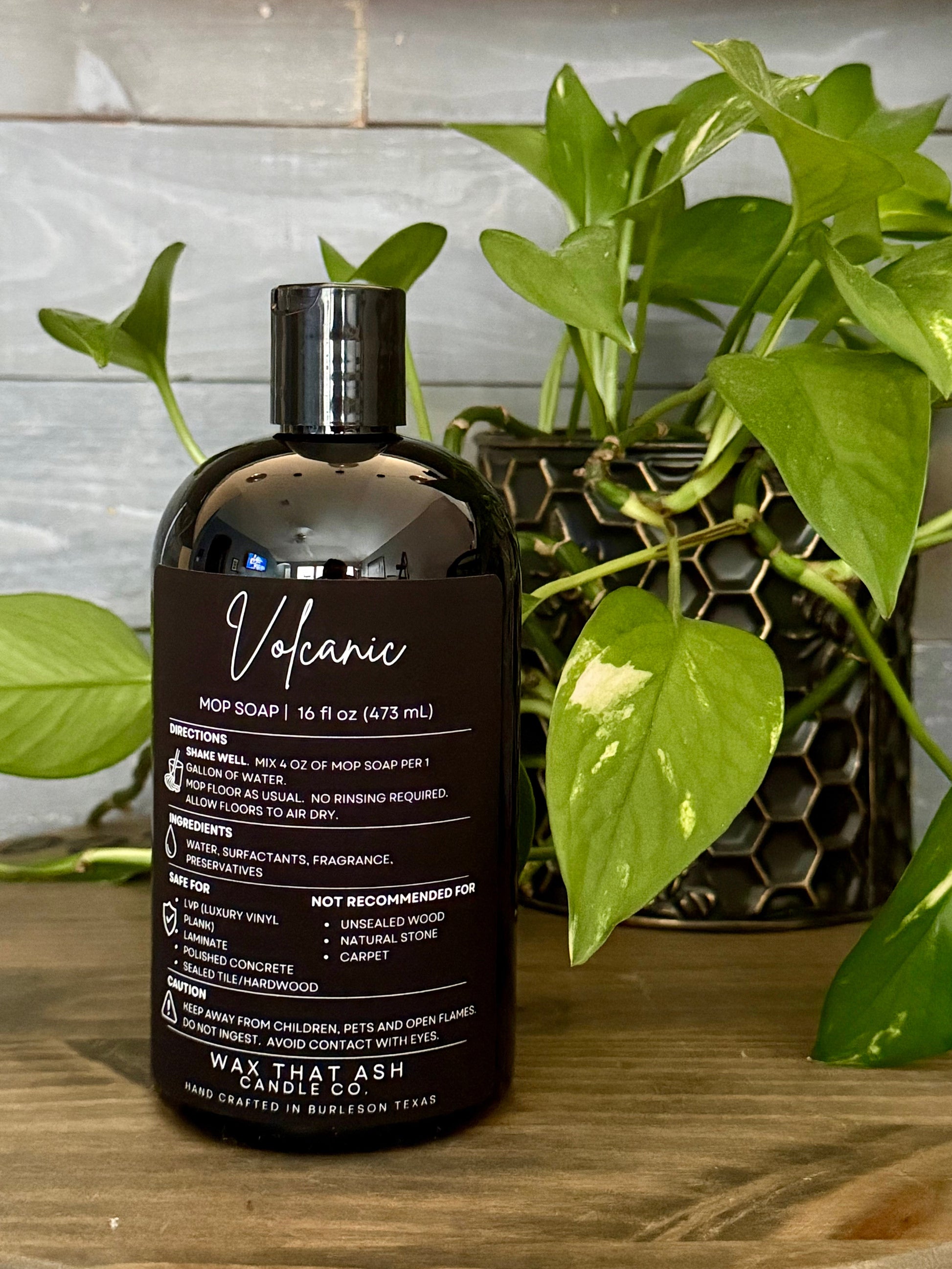 A bottle of Volcano scented hot soap by That Ash Candle Co. placed on a wooden surface next to a green pothos plant, with a gray tiled wall in the background.