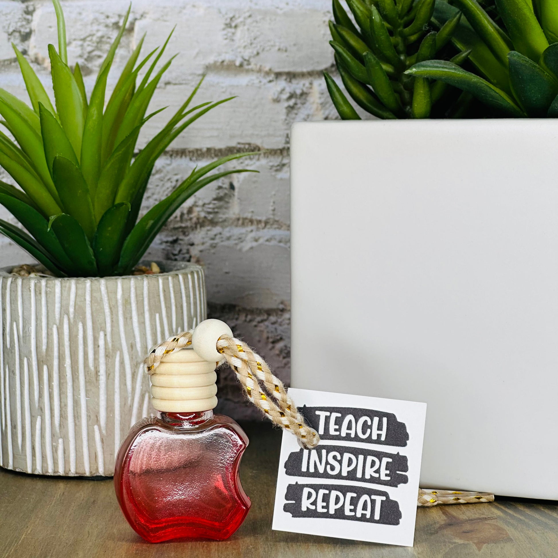 Red car air freshener with a tag reading 'Teach Inspire Repeat' on a wooden surface with plants in the background.