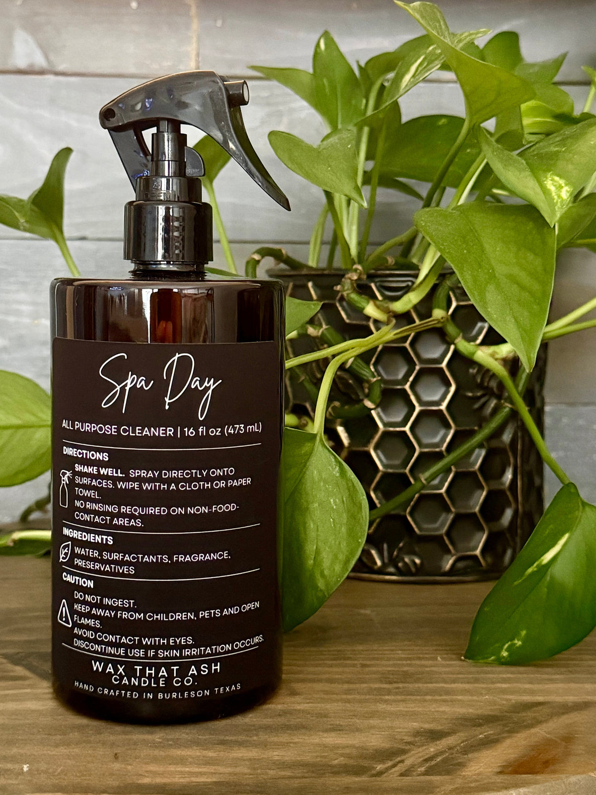 A bottle of Wax That Ash Spa Day all-purpose cleaner with a black spray trigger top, placed on a wooden surface next to a green potted plant.