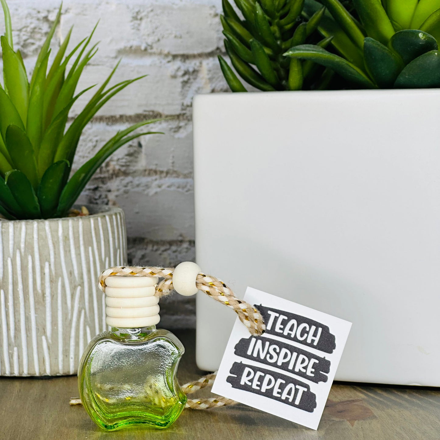 Small green bottle with a tag reading 'Teach Inspire Repeat' next to potted plants on a wooden surface.