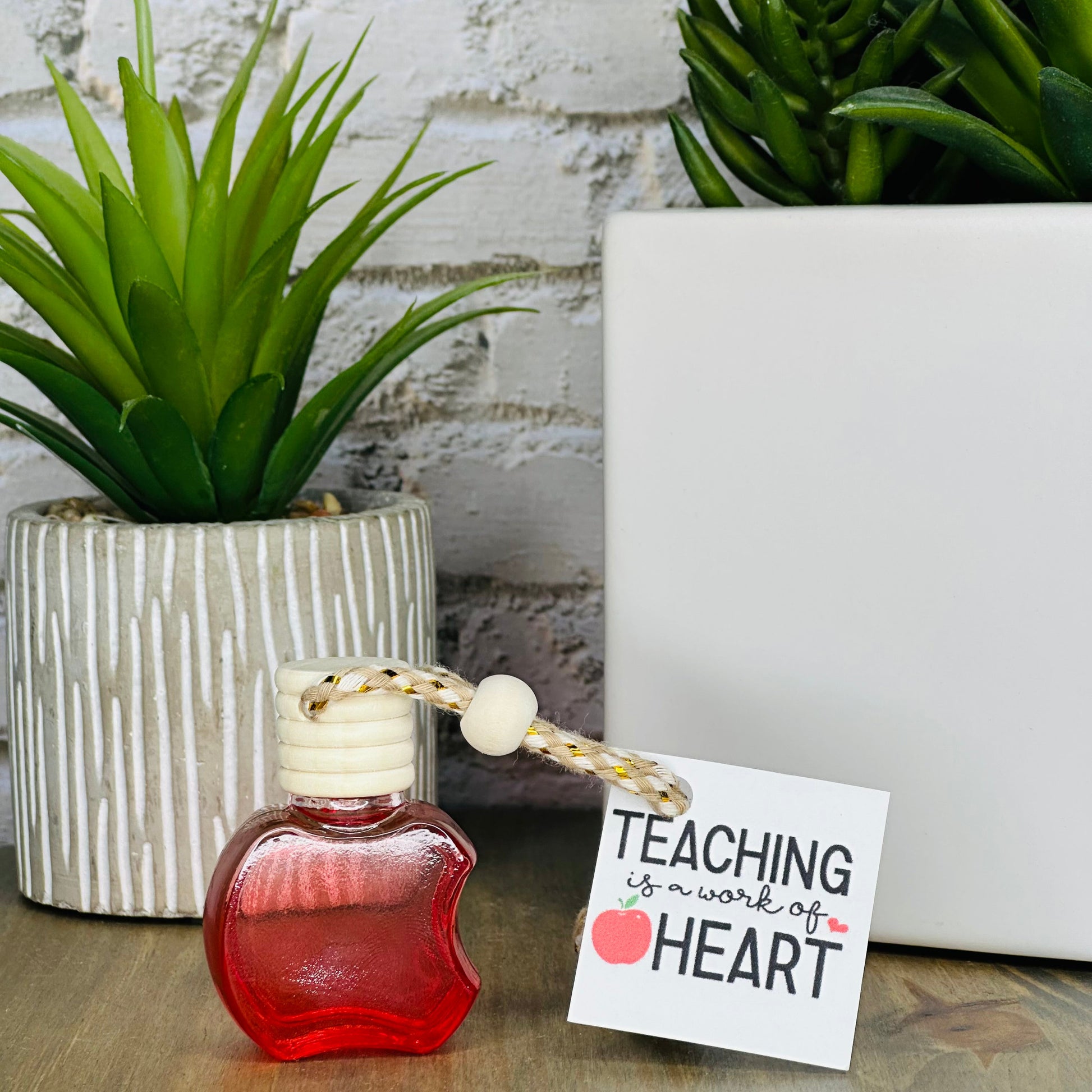 Small red bottle with a white cap on a wooden surface, next to potted plants and a sign that reads 'Teaching is a work of the heart'.